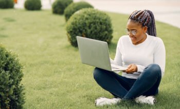 Young attractive dark-skinned college student. Lady sitting on the lawn at campus on sunny day. Girl working on her thesis using laptop computer.