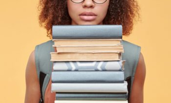 Portrait of scared black student girl in eyeglasses confused with workload holding heap of textbooks against yellow background